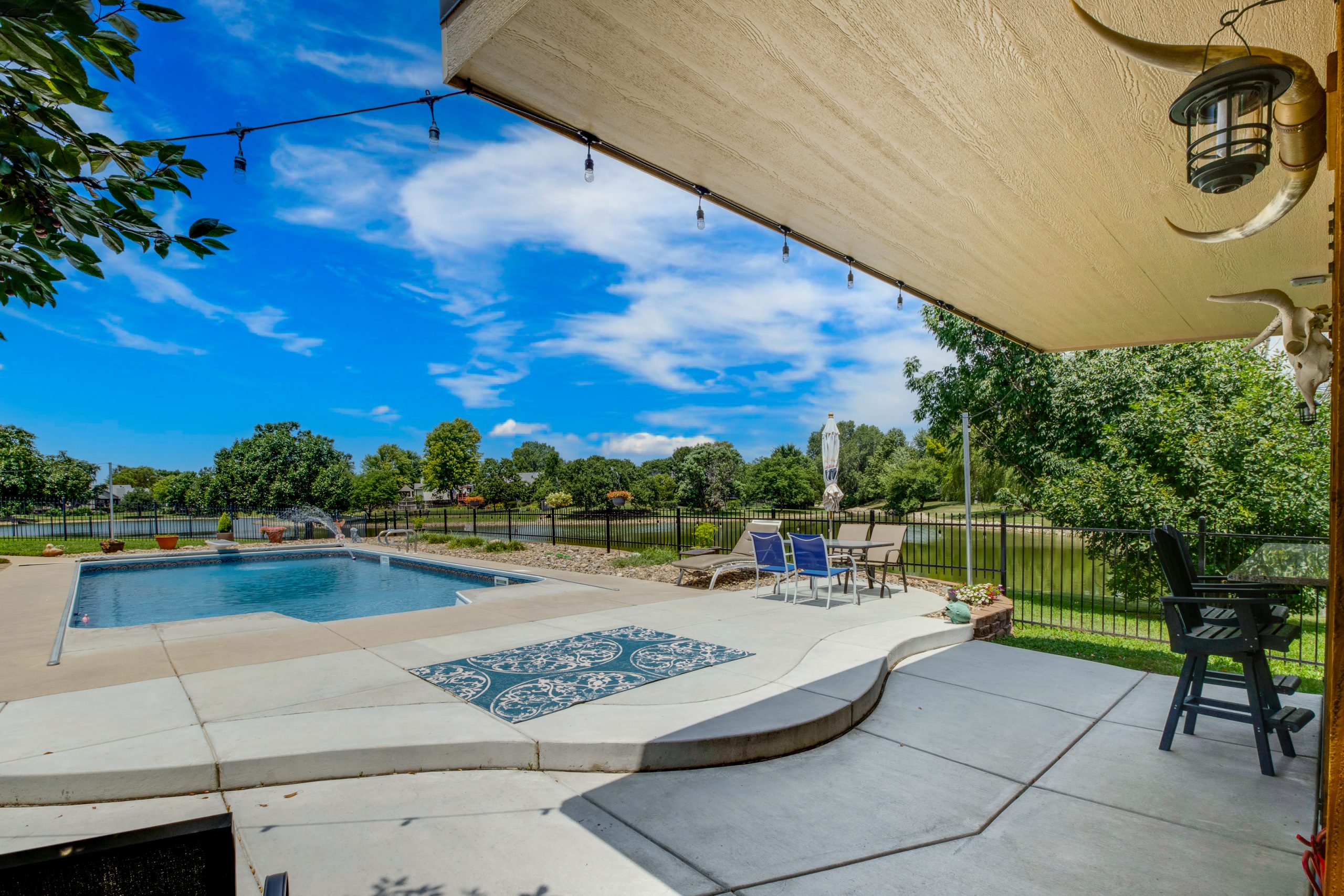 Backyard pool area with blue water, concrete deck, and string lights overhead; a fenced yard with trees beyond.