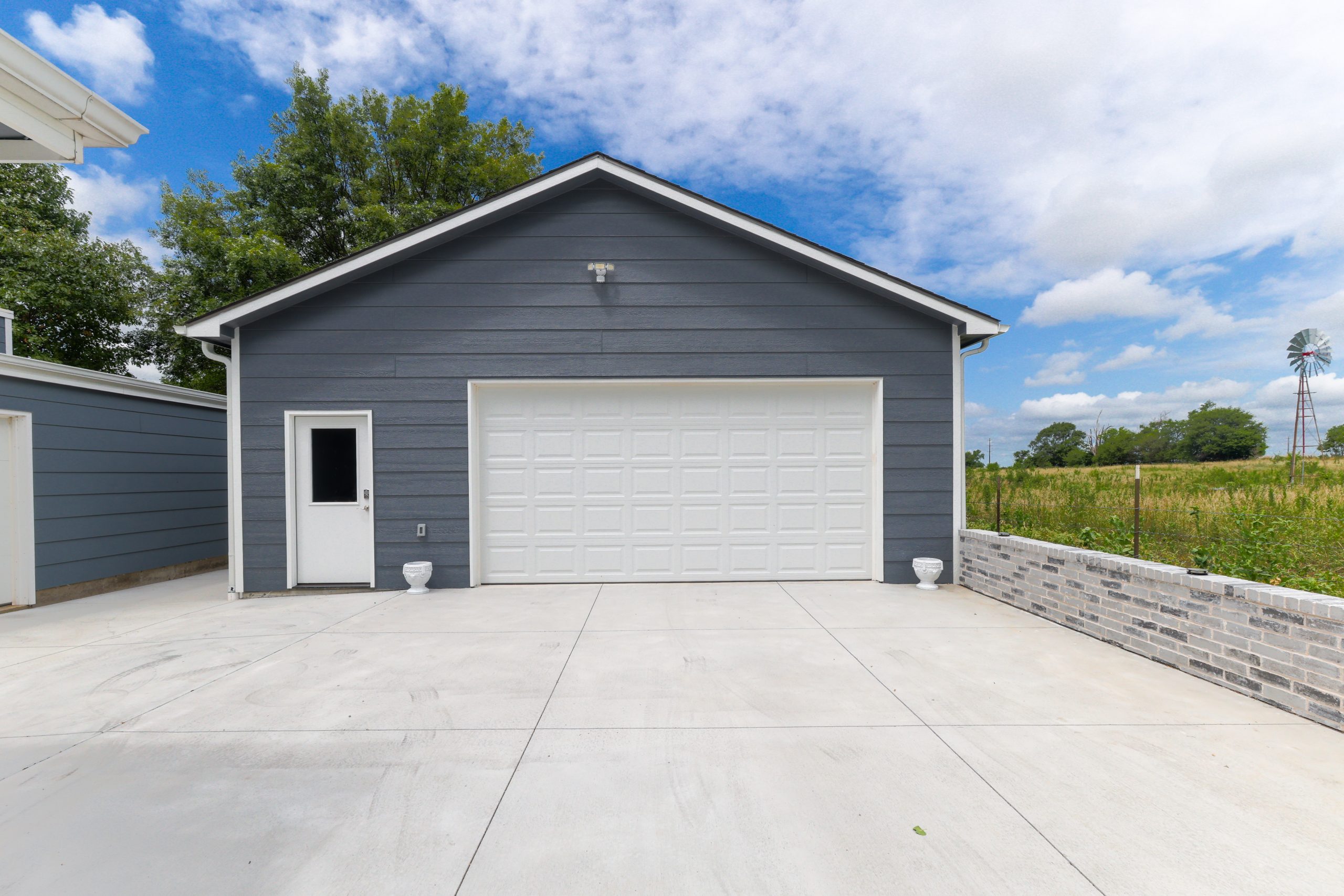 Front view of a blue-gray garage with a white sectional door and a small side door, driveway in foreground and a windmill in the field beyond.
