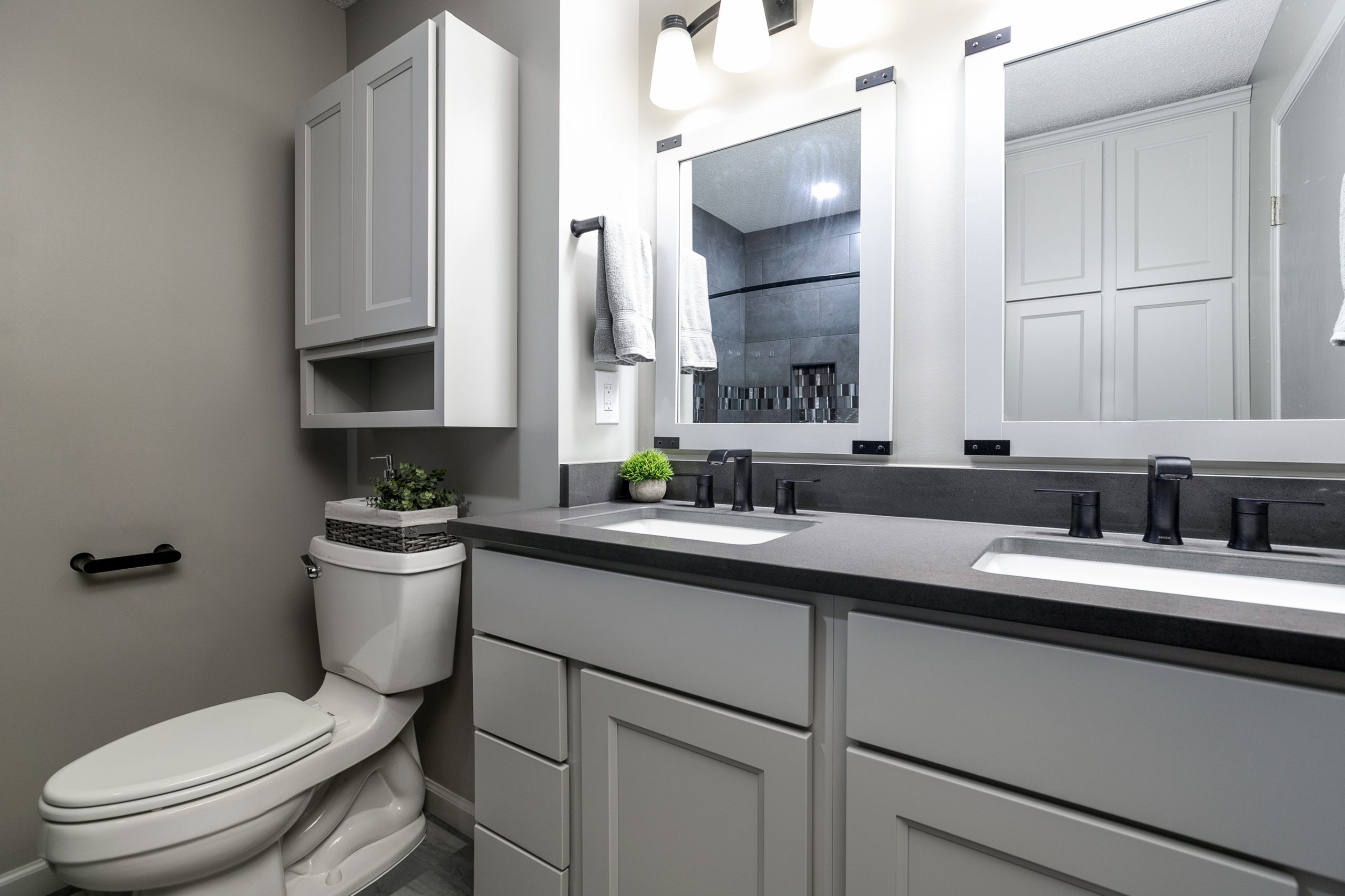 Modern gray bathroom with a dual-sink vanity, black fixtures, large framed mirrors, gray cabinetry, and a toilet with a small plant on the counter.