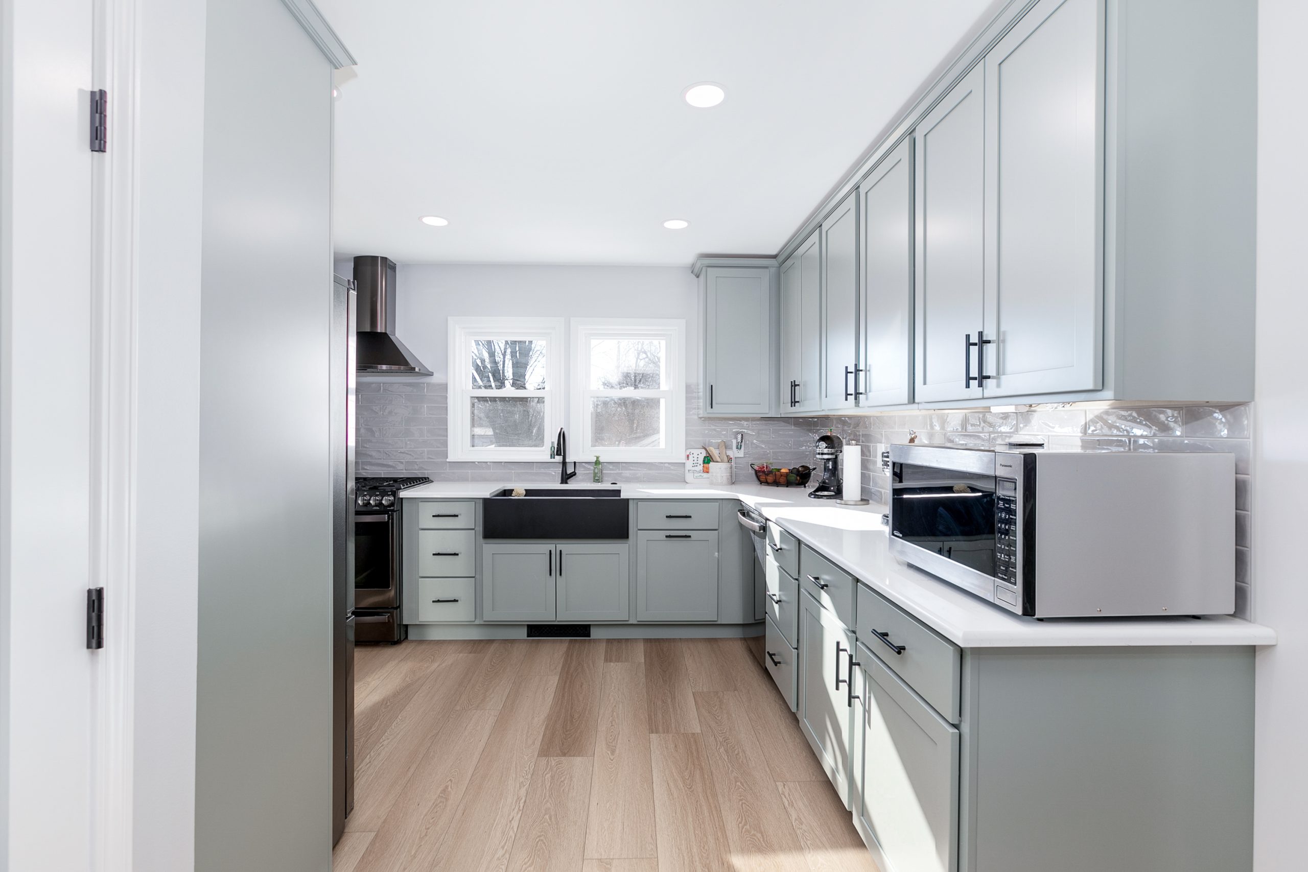 Modern kitchen with light gray cabinets, stainless steel appliances, and a wooden floor; ample natural light from two windows above the sink.