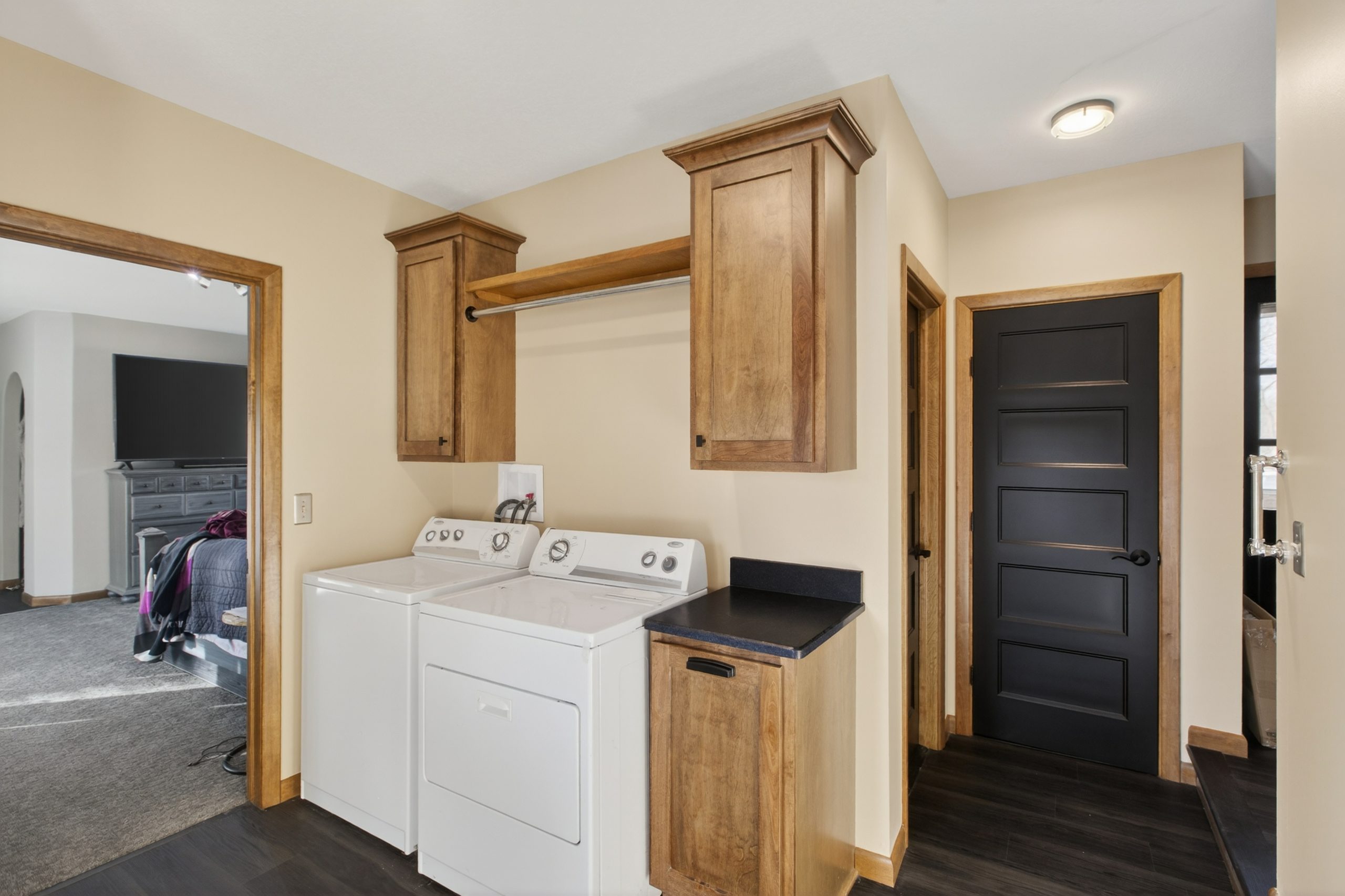 Laundry area with a washer and dryer under wooden cabinets and a hanging rod, in a beige-walled hallway.