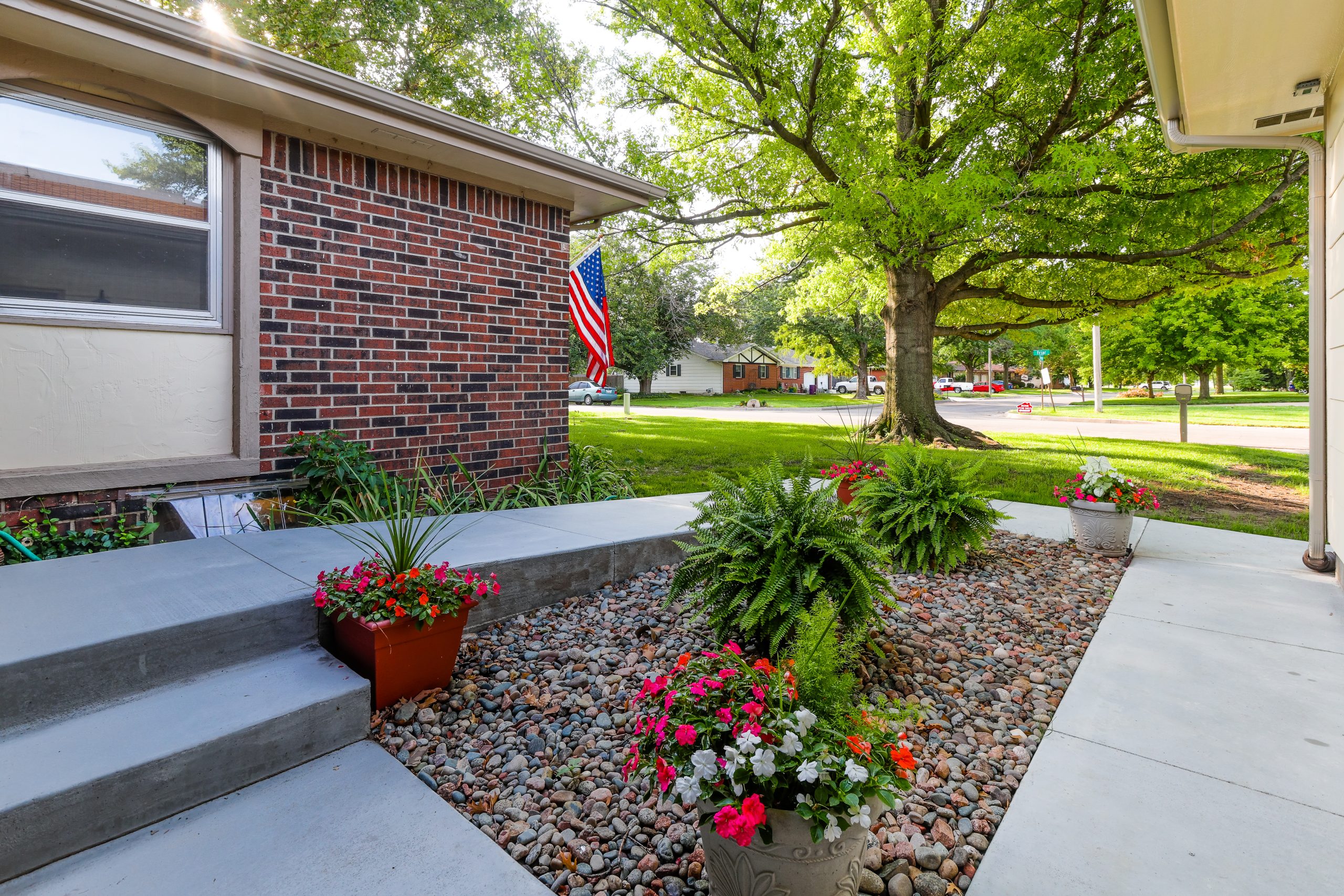 Brick house front yard with an American flag, flower pots, and a large leafy tree over a pebbled garden area by a suburban street.