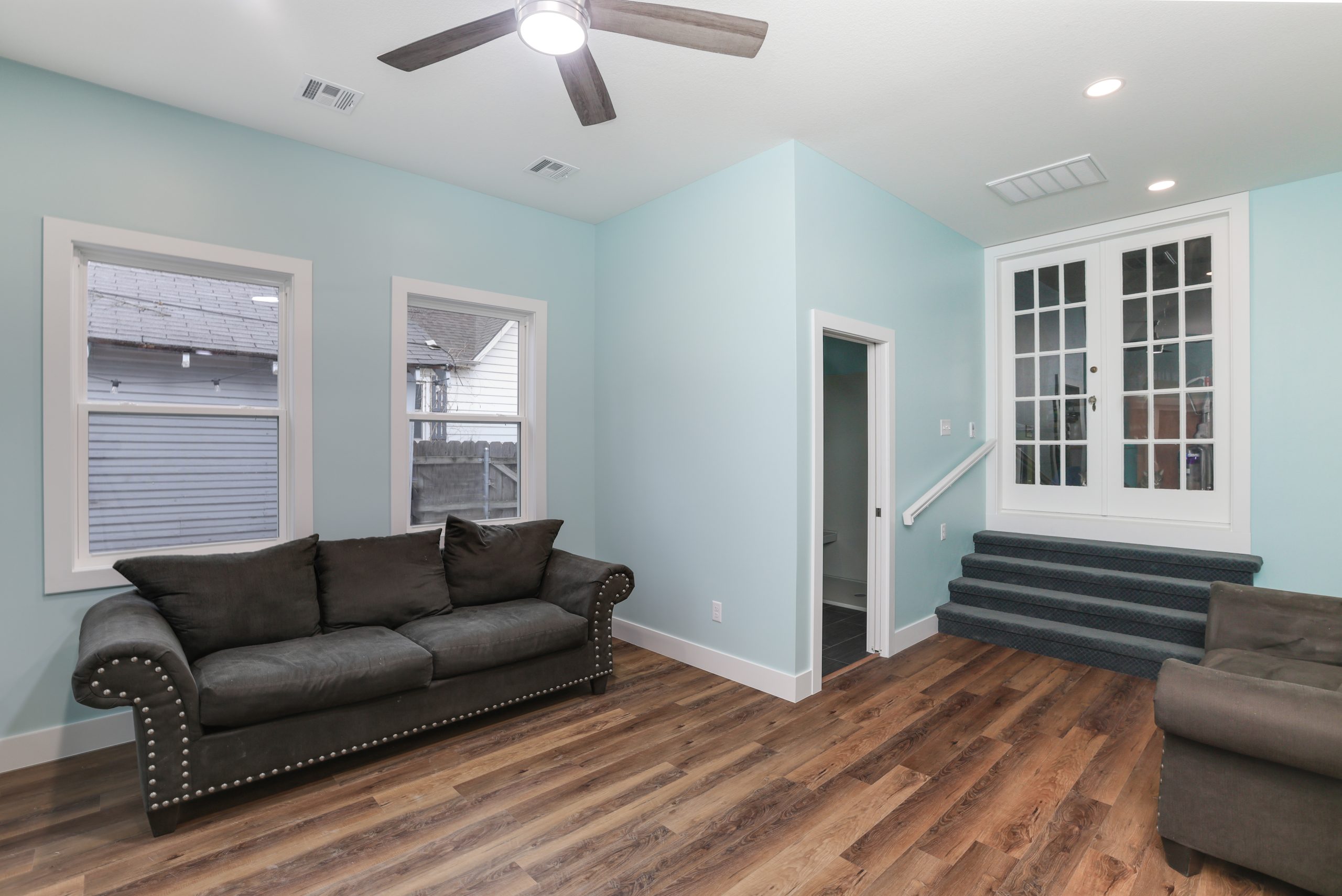 Living room with pale blue walls, dark gray upholstered sofa, and white-framed windows; carpeted stairs and white French doors to the right.