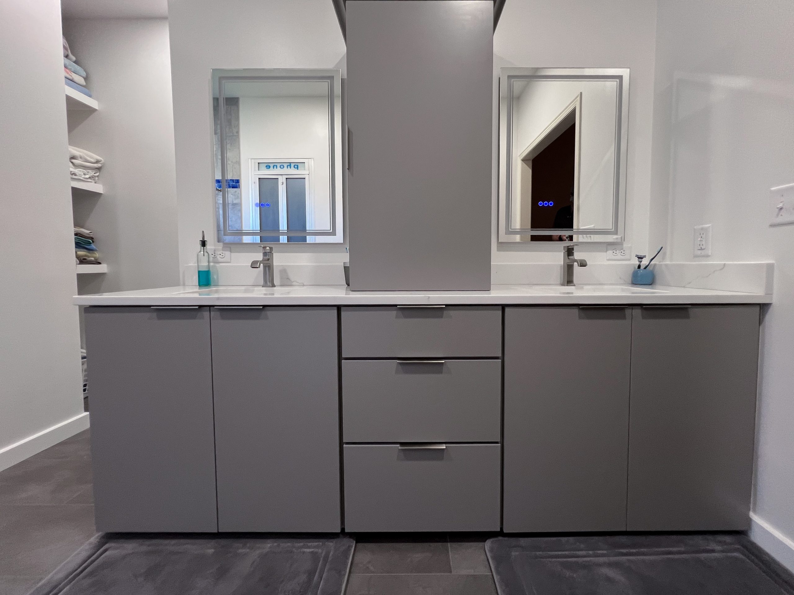 Double-sink bathroom vanity with two mirrors, gray cabinets, and a central column; towel shelves mounted to the left.