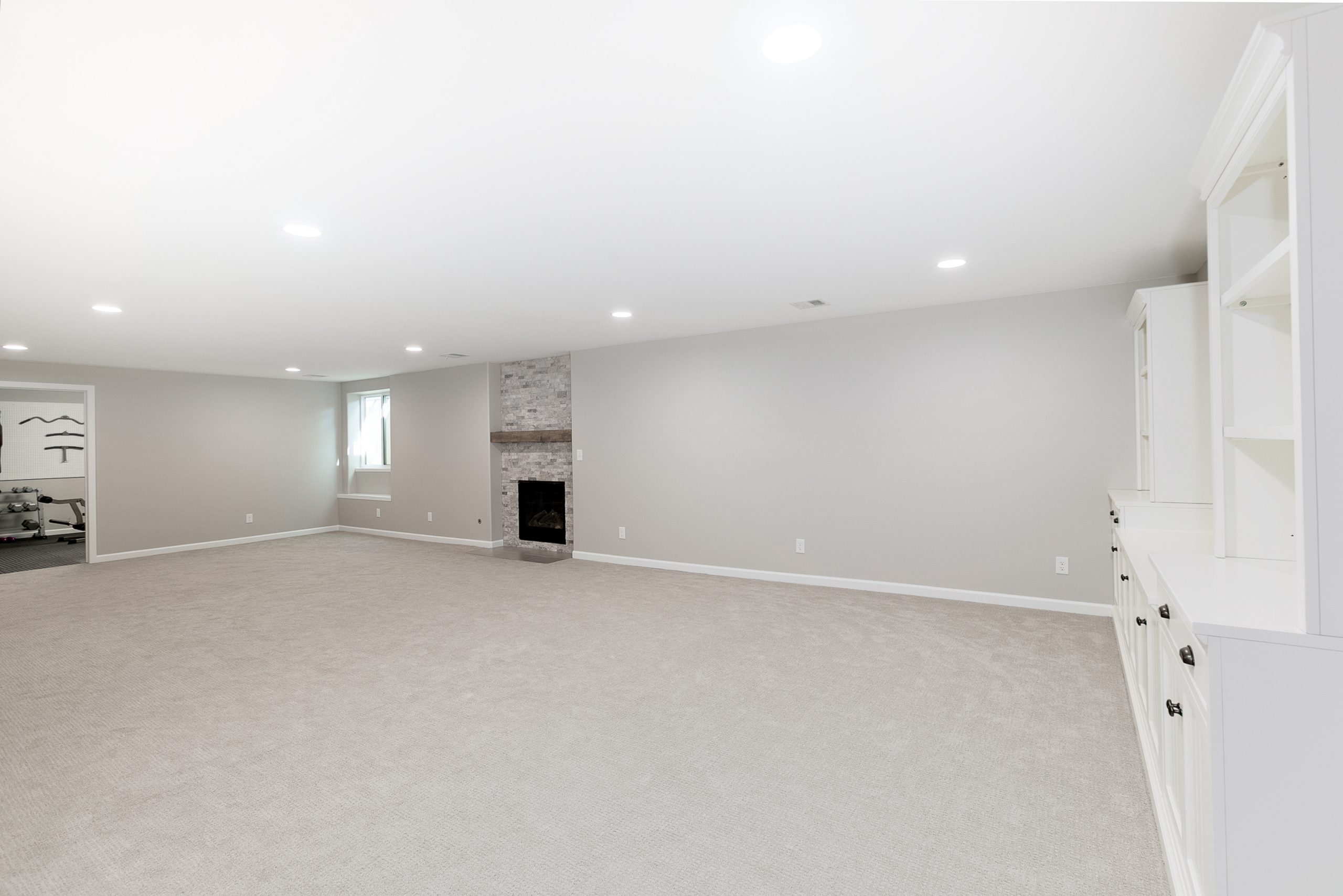 Spacious finished basement with white built-in cabinets along the right wall, a stone fireplace in the center, and a beige carpeted floor. A small exercise area is visible to the left.
