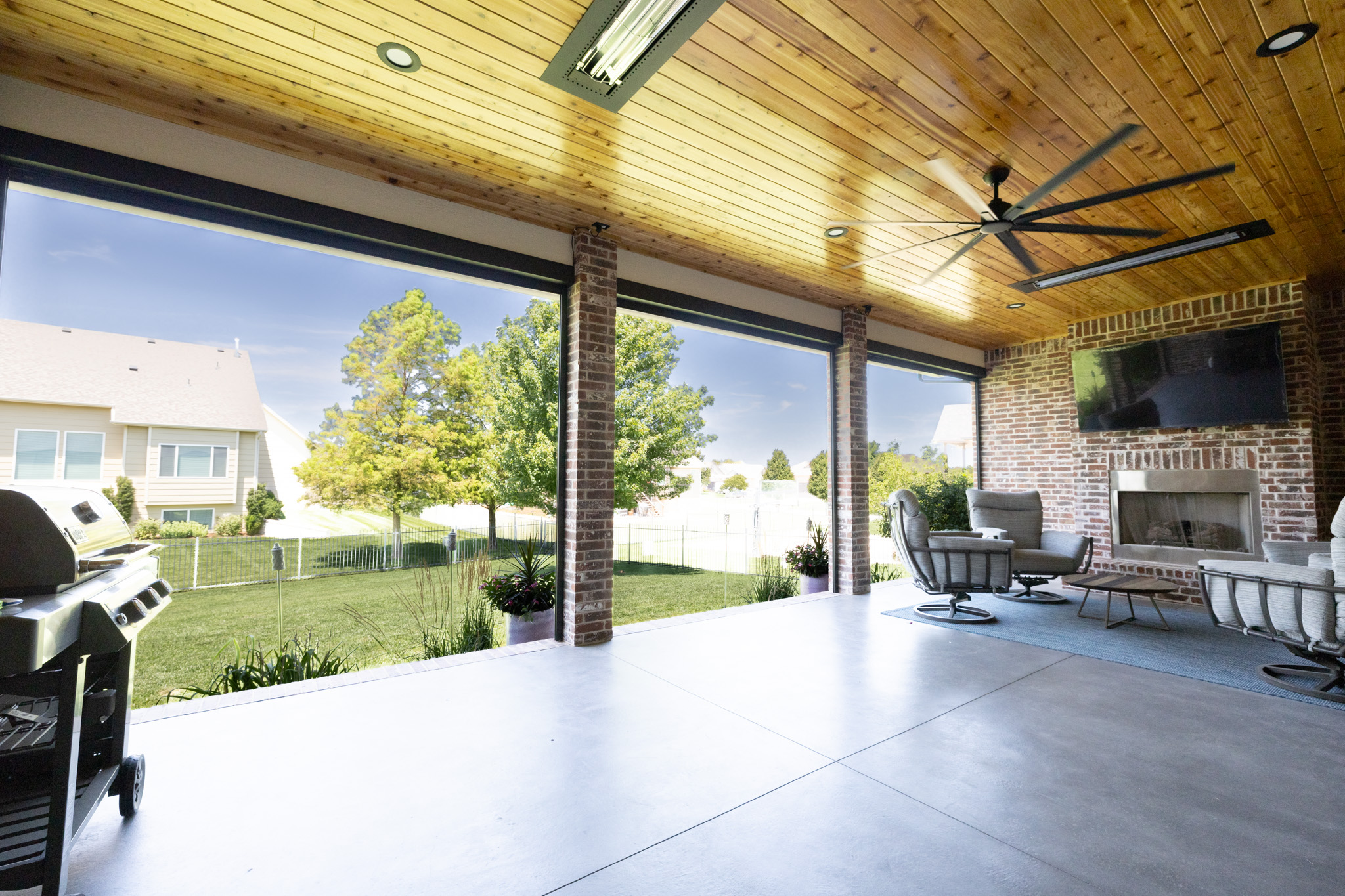 Covered outdoor patio with brick columns, wooden ceiling, and ceiling fan overlooking a grassy backyard and houses beyond. (informative)