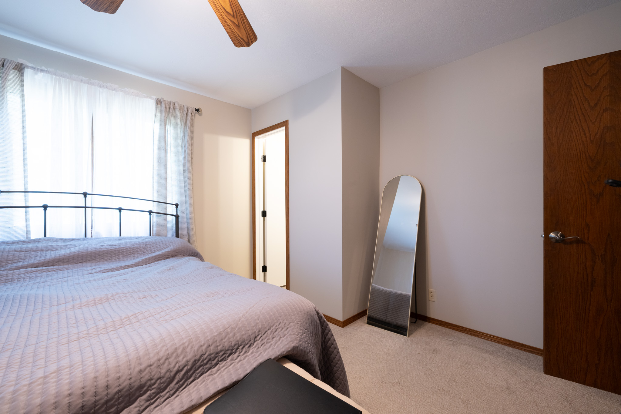 Bedroom with a metal-framed bed and gray quilted bedding, near a large window with sheer white curtains