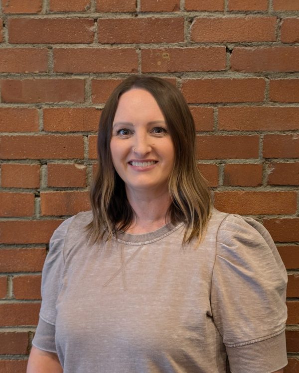 Smiling woman with shoulder-length brown hair and a nose ring standing in front of a red brick wall wearing a light gray top下-side view
