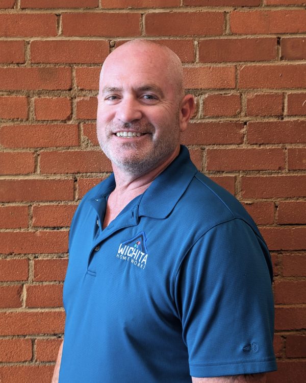 Portrait of a smiling man in a blue polo shirt with the Wichita Home Works logo, standing in front of a red brick wall.